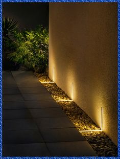 an illuminated pathway with rocks and plants on the side of a building at night time