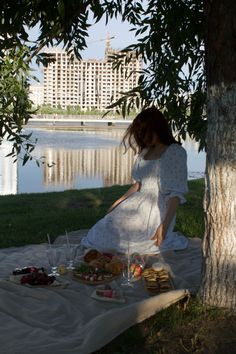 a woman in white dress sitting next to a tree with food on the ground near water