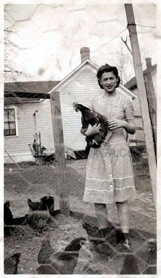 an old black and white photo of a woman holding a chicken in front of a house