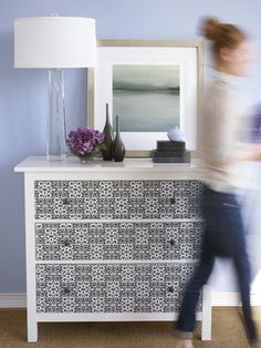 a woman is walking past a dresser with flowers on it and a framed photo in the background