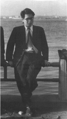 a man in a suit and tie standing next to a bench near the water with his hands on his hips