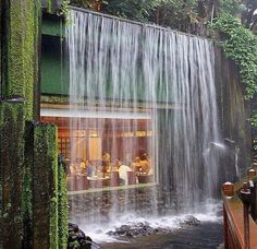 people are sitting at tables in front of a waterfall that is falling down the side of a building