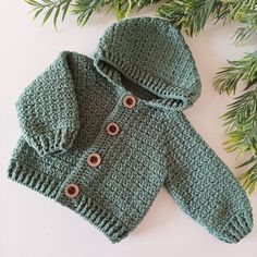 a crocheted green sweater with buttons on the hood and sleeves is laying next to some pine branches