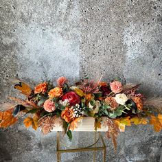 an arrangement of flowers and leaves on a gold stand against a gray wall with concrete flooring