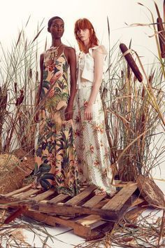 two women standing next to each other in front of tall grass and reeds,