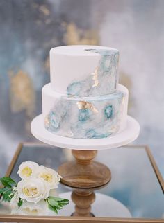 a marbled three tiered cake with gold leaf accents on a glass table next to white flowers