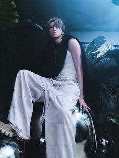 a man in white pants and black shirt sitting on top of a metal ball with plants behind him