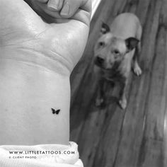 a small butterfly tattoo on the wrist of a person with a dog behind it,