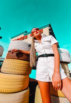 a woman in white shirt and shorts standing next to large stack of tires with sunglasses on