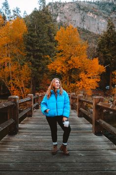 a woman standing on a wooden bridge in front of trees with yellow leaves and mountains behind her