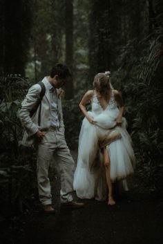 a man and woman dressed in white are walking through the woods with one holding on to her dress