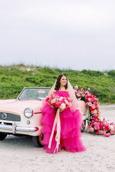 a woman in a pink dress standing next to an old car with flowers on it