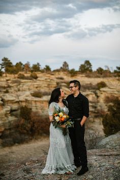 a man and woman standing next to each other on top of a rocky hill with trees in the background