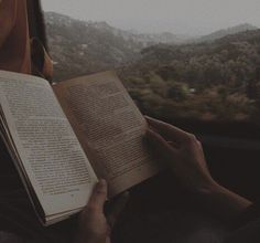 a person is reading a book while looking out the window at mountains and trees in the distance