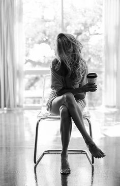 a woman sitting in a chair holding a coffee cup and looking out the window with her legs crossed