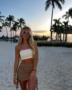 a beautiful young woman standing on top of a sandy beach next to palm trees at sunset