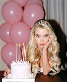 a blonde woman sitting in front of a cake with candles on it and pink balloons behind her