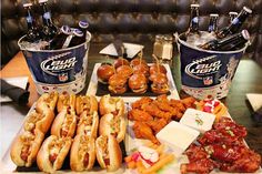 two buckets filled with beer bottles next to some food on a table and another tray full of hot dogs