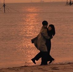 two people are walking on the beach near the water at sunset, with one person holding his arm around the other's neck