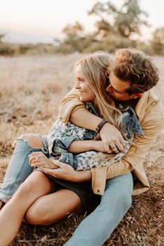 a man and woman sitting on the ground hugging each other in an open field with trees in the background