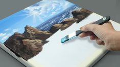 a hand is holding a paintbrush and painting a landscape on a piece of paper