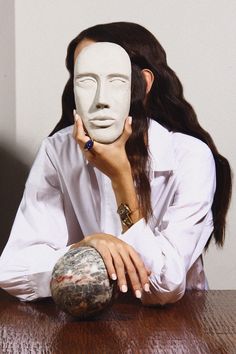 a woman sitting at a table with a white mask on her face and a rock in front of her