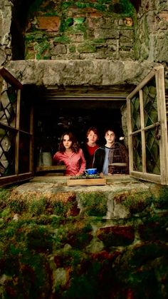 three people sitting in an open window on the side of a stone building with moss growing all over it