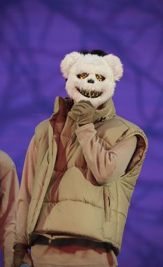 a person in a bear mask and jacket on stage with his hand up to his face