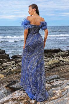 a woman in a blue dress standing on rocks next to the ocean and looking off into the distance