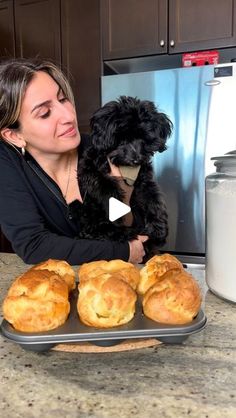 a woman holding a black dog in her arms next to some baked goods on a tray