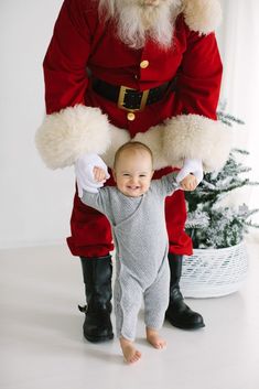 a baby standing next to santa clause in front of a christmas tree