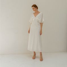 a woman in a white dress stands against a wall and looks at the camera while wearing sandals