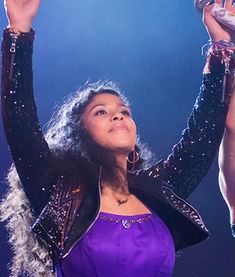 a woman in a purple dress holding her hands up to the sky while performing on stage