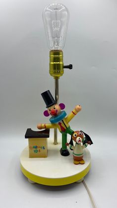 Bright and cheerful Vintage nursery lamp with a wide eyed, pink cheeked, red nosed clown wearing a tall black top hat and yellow shirt and green pants, arms spread wide and leaning on his balloon stand (which also serves as a night light)  Tested. Switch operated to turn on just the night light (behind the balloon stand), just the regular light (shown here w/ a 100 watt bulb--the only one I had at time of photo), or with both lights glowing. This is in good vintage condition with no chipped pain Circus Lamp, Balloon Stand, Nursery Lamp, Balloon Stands, Circus Clown, Yellow Shirt, Vintage Nursery, Balloon Wall, Red Nose