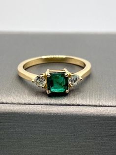 an emerald and diamond three stone ring on top of a silver box with a book in the background