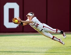 a female baseball player catching a ball in mid air with her glove on the ground