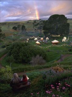 two people are sitting on a bench looking at a rainbow in the sky above them