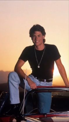 a man sitting on the hood of a car with his hands in his pockets and smiling