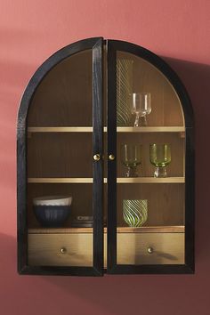 an arched glass cabinet with wine glasses on it's shelves, against a pink wall