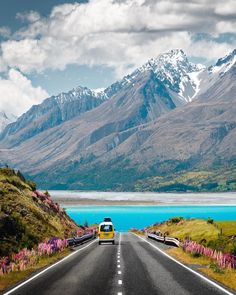 a van is driving down the road in front of some mountains and blue water with pink flowers