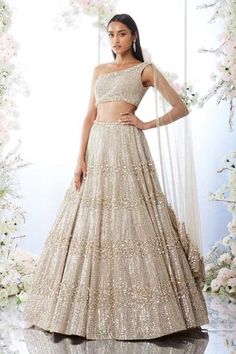 a woman standing in front of a floral backdrop wearing a gold and white lehenga