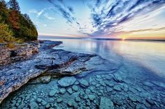 the sun is setting over an ocean with rocky shorelines and trees in the background