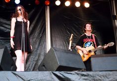 two people standing on stage with guitars and microphones in front of them, one playing the guitar