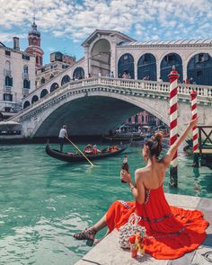 a woman in an orange dress sitting on the edge of a bridge with gondola passing by