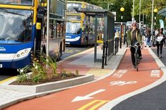 people walking and riding bikes on the sidewalk next to buses parked in front of them