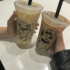 two people holding up cups with drinks in them sitting on a white table top together