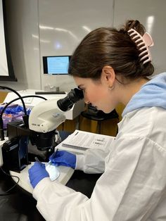 a woman in white coat and blue gloves looking through a microscope at something on the table