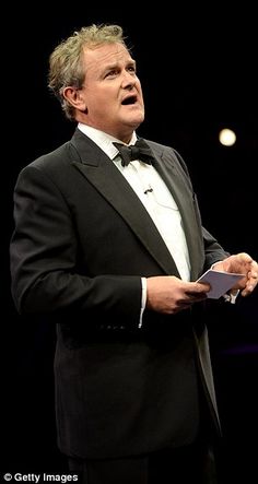 a man in a tuxedo is speaking on stage with his hands folded open