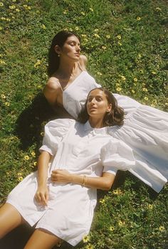 two women laying in the grass with their arms around each other, one wearing a white dress