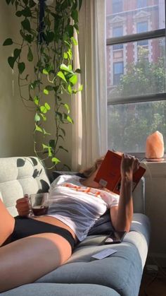 a woman laying on a couch reading a book and holding a drink in her hand